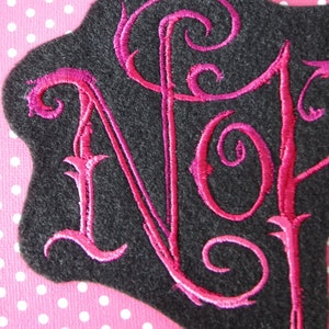 May include: A black felt patch with the letters "NJ" embroidered in pink cursive.