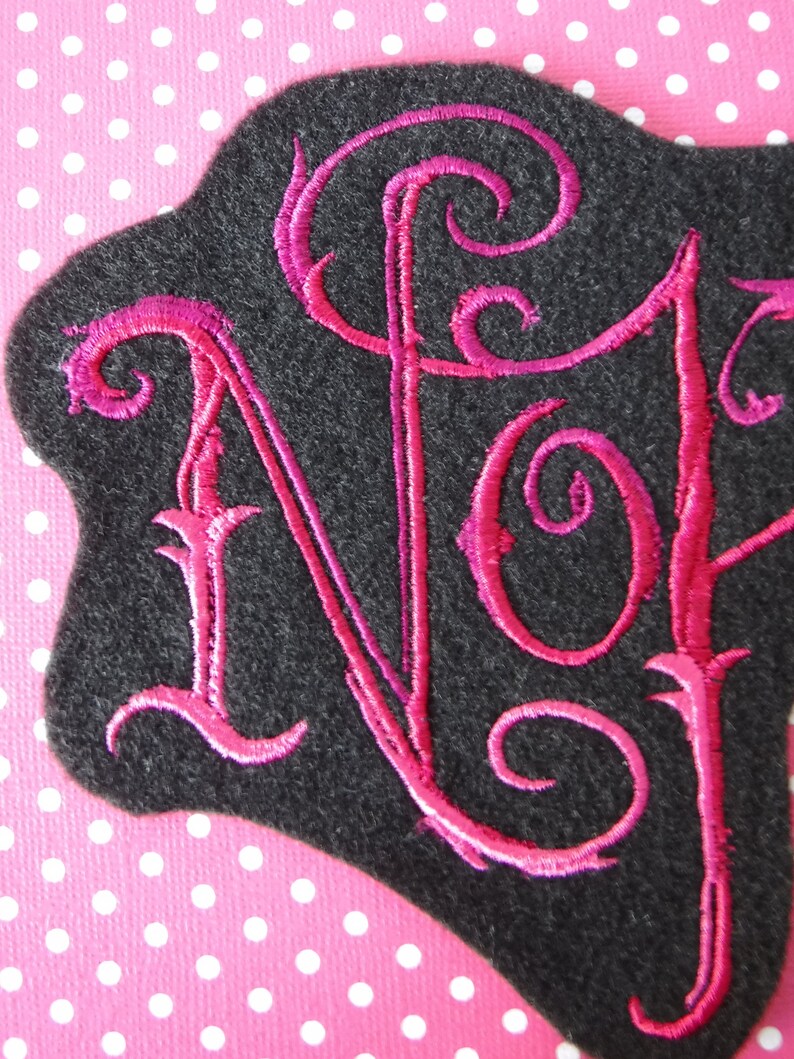 May include: A black felt patch with the letters "NJ" embroidered in pink cursive.