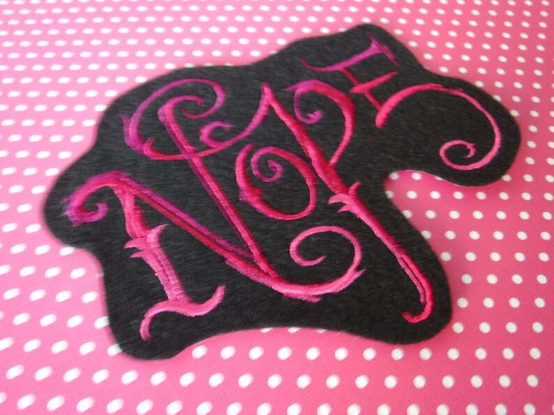 May include: A black felt patch with the word "NOPE" embroidered in pink thread. The letters are in a stylized font with swirls.