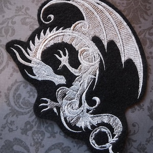 May include: A white embroidered dragon design on a black fabric patch. The dragon is curled up with its wings spread out.