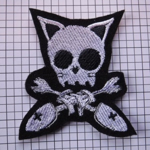 May include: Black and white embroidered patch of a skull with cat ears and crossbones. The skull is white with black eyes and a black nose. The crossbones are white and are behind the skull.