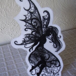 May include: Black embroidered fairy silhouette with intricate lace wings and a flowing dress. The fairy is depicted against a white background.