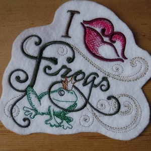 May include: Embroidered patch with the text "I love frogs" in a cursive font. A green frog with a gold crown is featured below the text. A red embroidered heart is above the text.