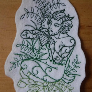 May include: White felt applique featuring a green embroidered illustration of a mythological figure playing a flute, surrounded by foliage. The figure has horns and a beard. The applique has an irregular, organic shape.
