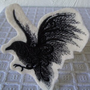 May include: Black embroidered patch of a bird with its wings spread out. The bird is on a white felt background.