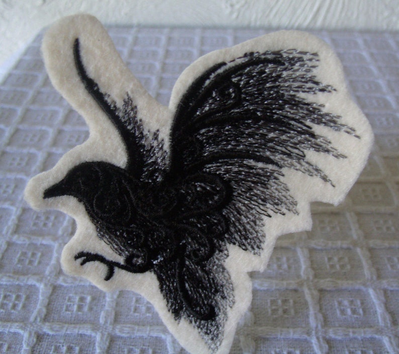 May include: Black embroidered patch of a bird with its wings spread out. The bird is on a white felt background.