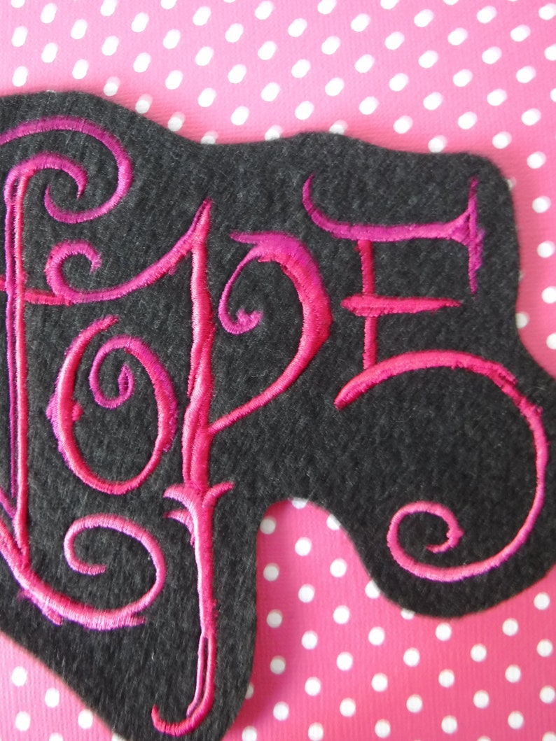 May include: A pink embroidered patch with the word "Hope" in a stylized font on a black felt background.