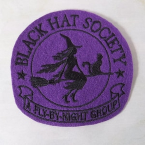 May include: A circular purple felt patch featuring black embroidery. The design shows a silhouette of a witch on a broomstick with a cat, surrounded by the text BLACK HAT SOCIETY and A FLY-BY-NIGHT GROUP, separated by two small black stars.