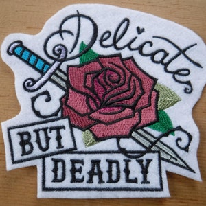 May include: Embroidered iron-on patch with a pink rose, two silver daggers, and green leaves. The text "Delicates But Deadly" is stitched in black thread.