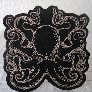 May include: Black felt patch with an embroidered white octopus design. The octopus has eight tentacles with swirling patterns.