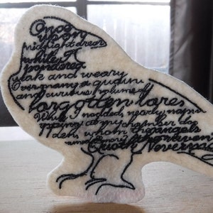 May include: White felt raven with black embroidery of the poem "The Raven" by Edgar Allan Poe. The poem is embroidered on the raven's body and wings.