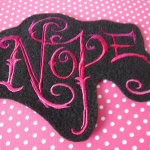 May include: A black felt patch with a pink embroidered word "NOPE" in a stylized font.