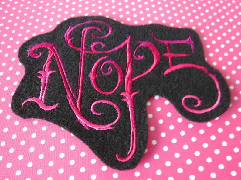May include: A black felt patch with a pink embroidered word "NOPE" in a stylized font.