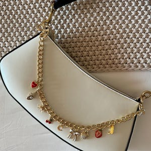 Op de afbeelding: Cr&egrave;mekleurige schoudertas met een zwarte band en gouden ketting. De ketting is versierd met diverse bedels, waaronder een rood hart, een parel, een kers, een strik, een schelp en een aardbei. De tas heeft een gebogen vorm en een zwarte rand.