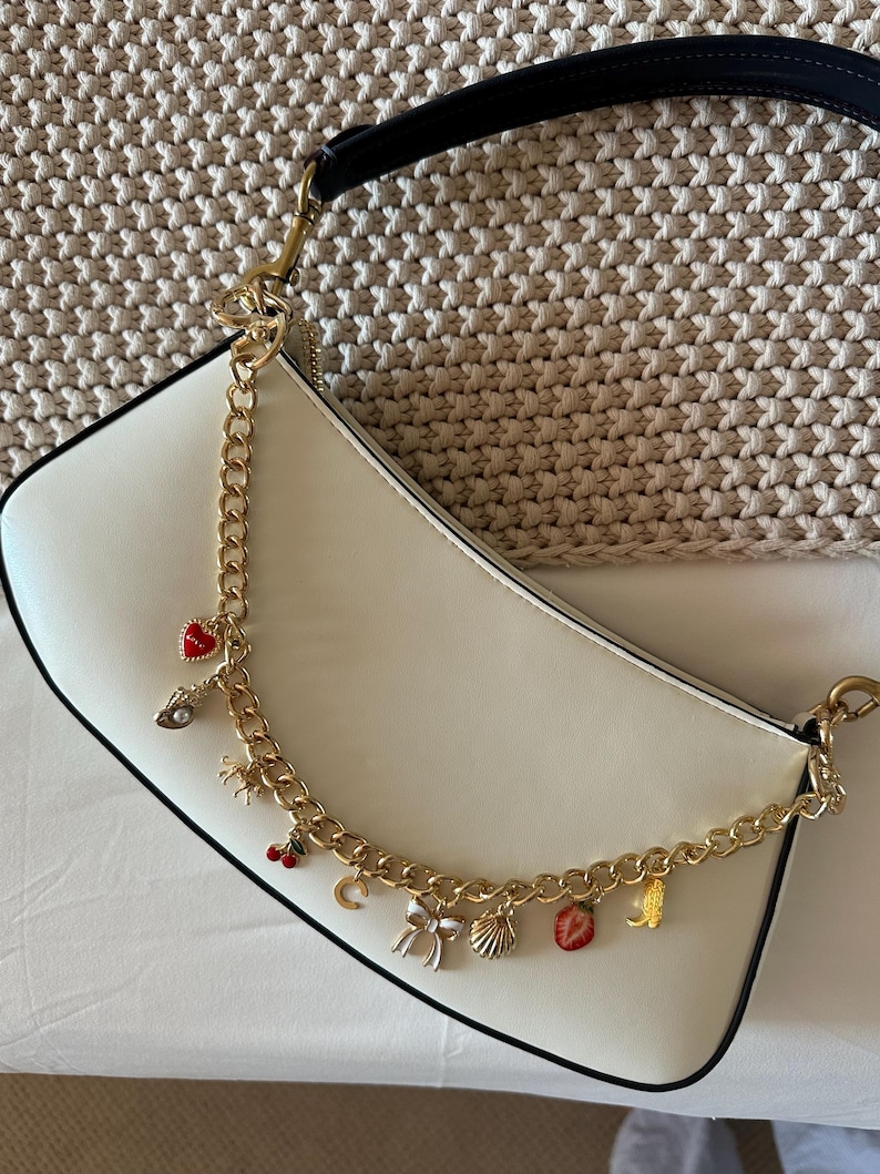 Op de afbeelding: Cr&egrave;mekleurige schoudertas met een zwarte band en gouden ketting. De ketting is versierd met diverse bedels, waaronder een rood hart, een parel, een kers, een strik, een schelp en een aardbei. De tas heeft een gebogen vorm en een zwarte rand.