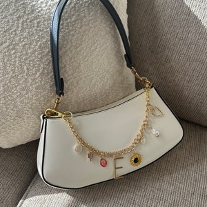 Op de afbeelding: Witte baguette-handtas met een zwart handvat en goudkleurige hardware. De tas heeft een gouden ketting met bedels, waaronder een aardbei, zonnebloem en de letter "E".