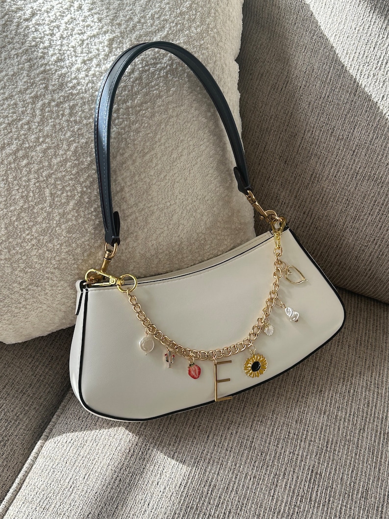 Op de afbeelding: Witte baguette-handtas met een zwart handvat en goudkleurige hardware. De tas heeft een gouden ketting met bedels, waaronder een aardbei, zonnebloem en de letter "E".