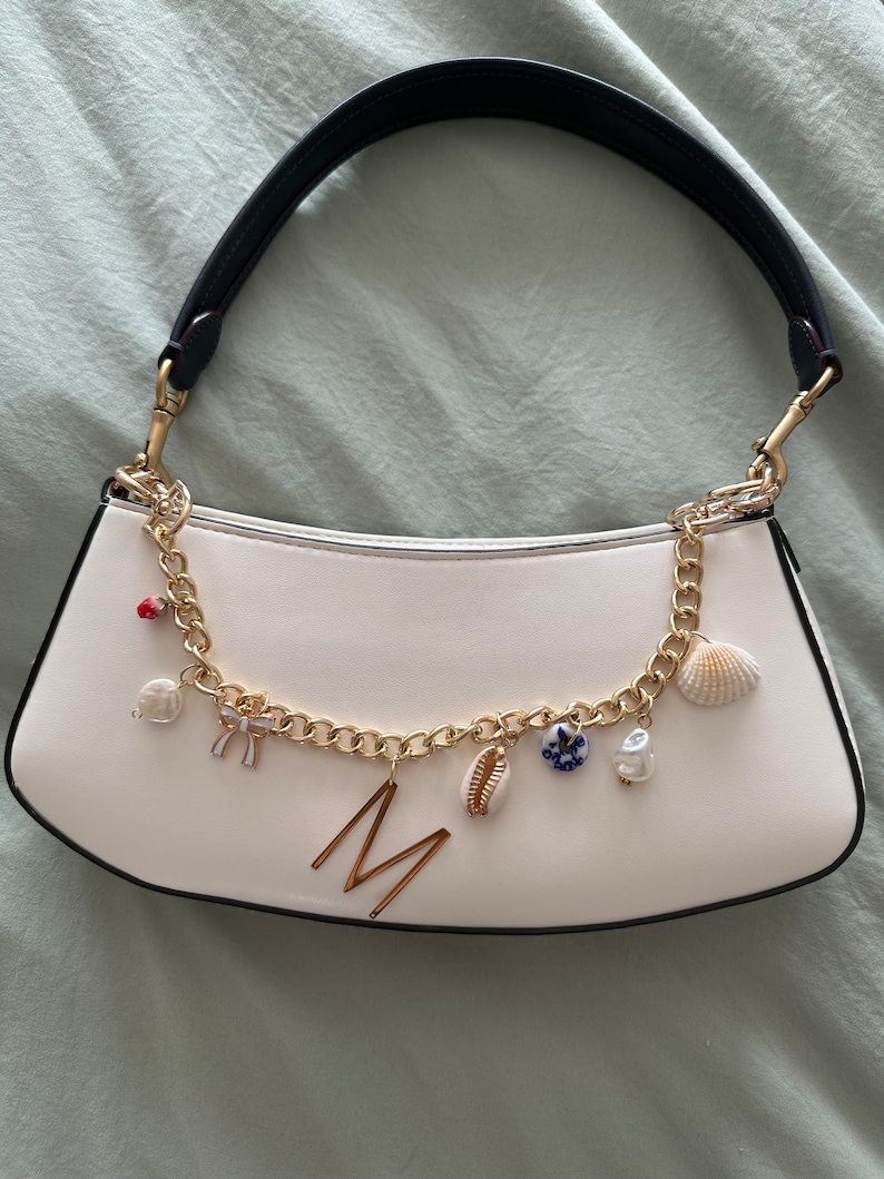 Op de afbeelding: Witte handtas met een zwarte handgreep en een goudkleurige ketting. De ketting is versierd met bedels, waaronder een zeeschelp, een parel en de letter "M". De tas heeft een gebogen vorm.