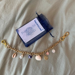 Op de afbeelding: Goudkleurige kettingarmband met diverse bedels, waaronder schelpen, een parel, een blauw-witte keramische kraal, een roze bloem en een zon. De armband heeft twee gouden sluitingen. Een klein blauw mesh zakje en een kaart met tekst zijn ook aanwezig.