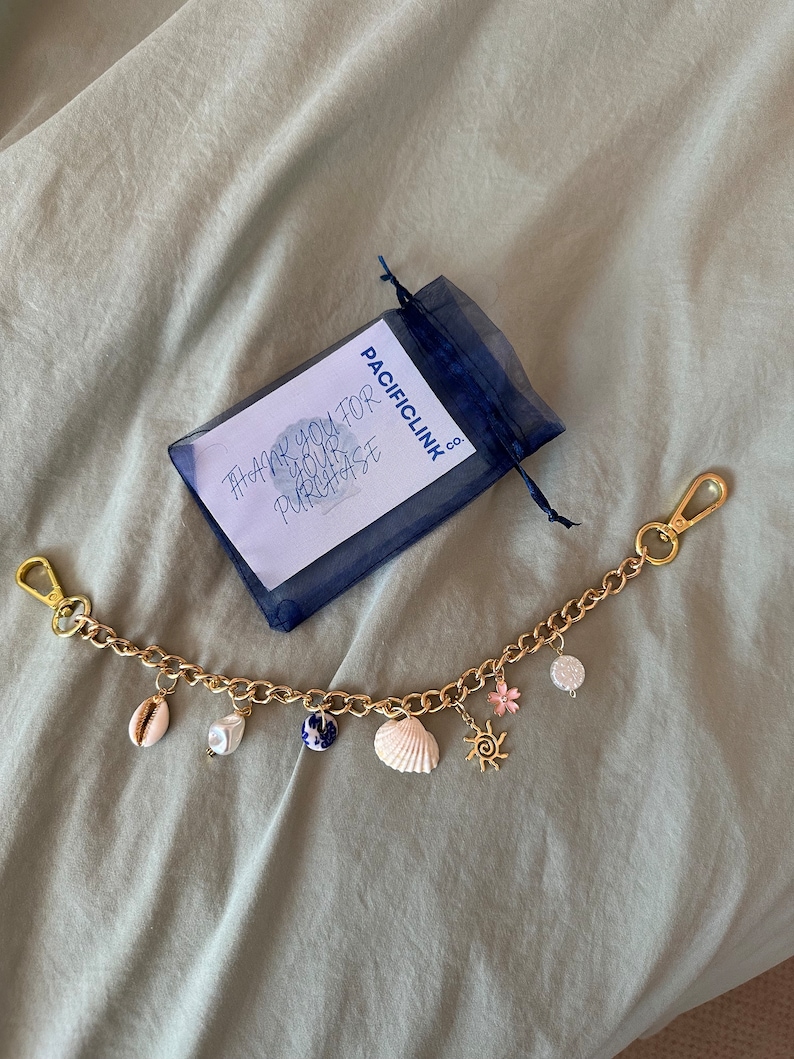 Op de afbeelding: Goudkleurige kettingarmband met diverse bedels, waaronder schelpen, een parel, een blauw-witte keramische kraal, een roze bloem en een zon. De armband heeft twee gouden sluitingen. Een klein blauw mesh zakje en een kaart met tekst zijn ook aanwezig.