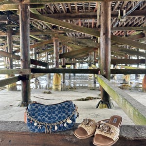 Op de afbeelding: Een denim handtas met een blauw monogrampatroon, een gouden ketting en bedels, naast geweven sandalen met gouden details. De items worden gepresenteerd op een houten oppervlak onder een pier.