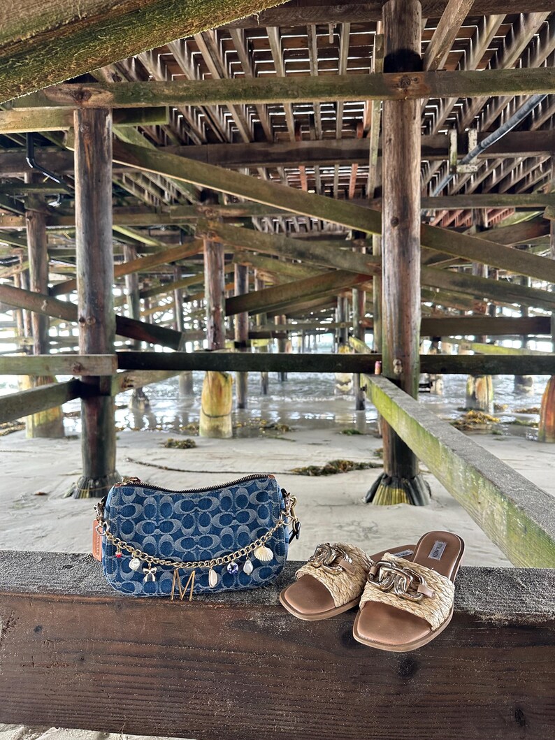 Op de afbeelding: Een denim handtas met een blauw monogrampatroon, een gouden ketting en bedels, naast geweven sandalen met gouden details. De items worden gepresenteerd op een houten oppervlak onder een pier.