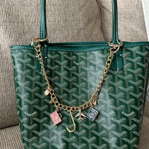 Op de afbeelding: Groene tote bag met witte stiksels en een gouden ketting. De tas heeft een herhalend patroon en een gouden ketting met reis-thema bedels, waaronder een koffer, een vliegtuig en een paspoort.
