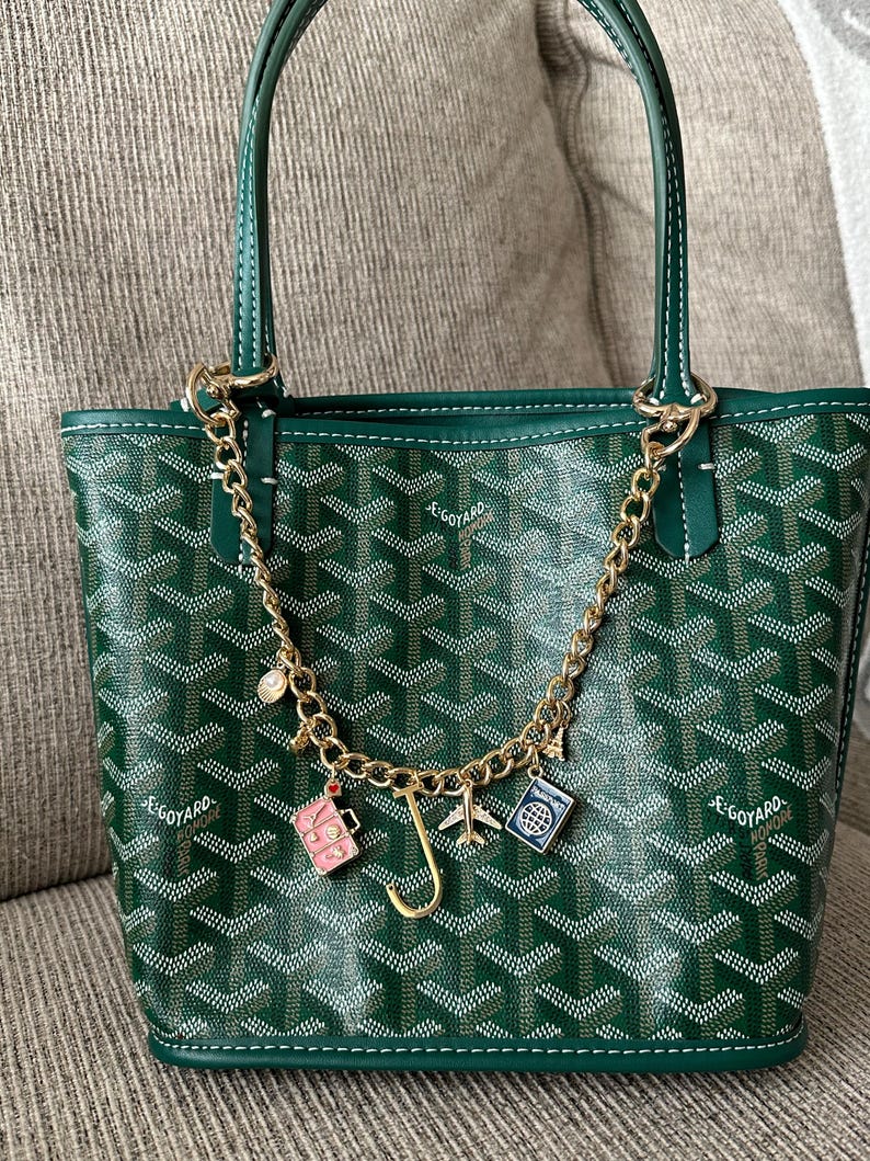 Op de afbeelding: Groene tote bag met witte stiksels en een gouden ketting. De tas heeft een herhalend patroon en een gouden ketting met reis-thema bedels, waaronder een koffer, een vliegtuig en een paspoort.