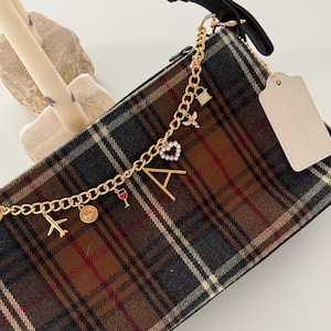 Op de afbeelding: Handtas met een bruin, grijs en rood ruitpatroon, met een gouden ketting met bedels. Bedels zijn onder meer een vliegtuig, een wijnglas, een hart en de letter "A". Het heeft een zwarte band en een Coach-label.