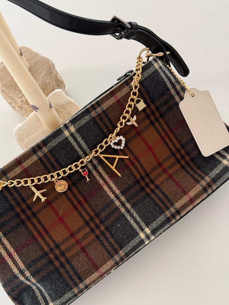 Op de afbeelding: Handtas met een bruin, grijs en rood ruitpatroon, met een gouden ketting met bedels. Bedels zijn onder meer een vliegtuig, een wijnglas, een hart en de letter "A". Het heeft een zwarte band en een Coach-label.
