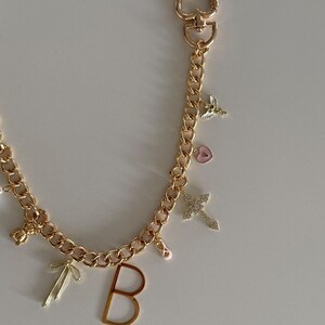 Op de afbeelding: Goudkleurige ketting met een hartvormige sluiting en diverse bedels. Bedels zijn onder meer een kruis, de letter "B", een strik, een balletschoen, een hart en een kleine engel. Op een witte achtergrond.