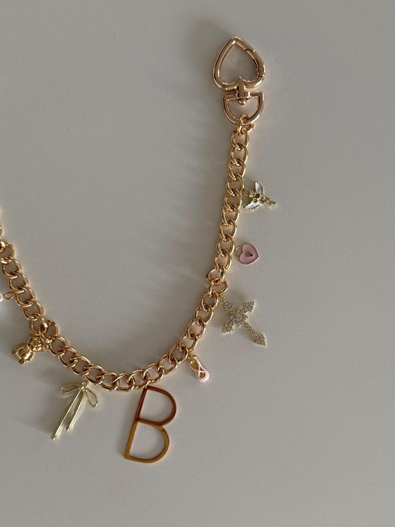 Op de afbeelding: Goudkleurige ketting met een hartvormige sluiting en diverse bedels. Bedels zijn onder meer een kruis, de letter "B", een strik, een balletschoen, een hart en een kleine engel. Op een witte achtergrond.