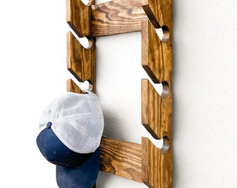 Puede incluir: Un perchero de madera para sombreros con un acabado de tinte oscuro, montado en una pared blanca. El perchero presenta múltiples ranuras anguladas diseñadas para sujetar gorras de béisbol. Una gorra azul y blanca cuelga de una de las ranuras.
