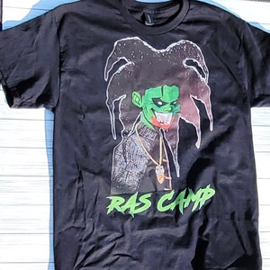 May include: Black t-shirt with a green and black cartoon character with dreadlocks and the text "RAS CAMP" in green.