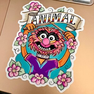 May include: A colorful illustration of Animal from the Muppets, holding drumsticks and a banner that says "ANIMAL". The illustration is surrounded by pink flowers.
