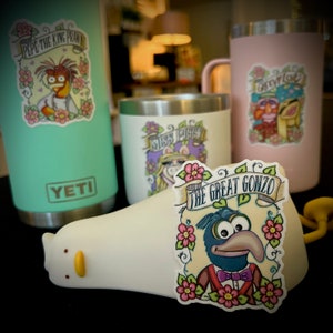 May include: Three stainless steel tumblers with colorful Muppet character stickers. The stickers feature Pepe the King Prawn, Miss Piggy, and The Great Gonzo. The tumblers are all Yeti brand.