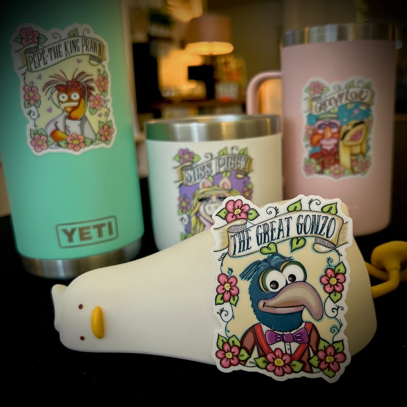 May include: Three stainless steel tumblers with colorful Muppet character stickers. The stickers feature Pepe the King Prawn, Miss Piggy, and The Great Gonzo. The tumblers are all Yeti brand.