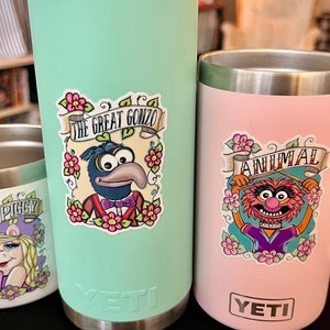 May include: Three stainless steel tumblers with colorful Muppet character stickers. The tumblers are in mint green, pink, and white. The stickers feature Miss Piggy, Animal, and Gonzo. The text on the stickers reads "Miss Piggy", "Animal", and "The Great Gonzo".