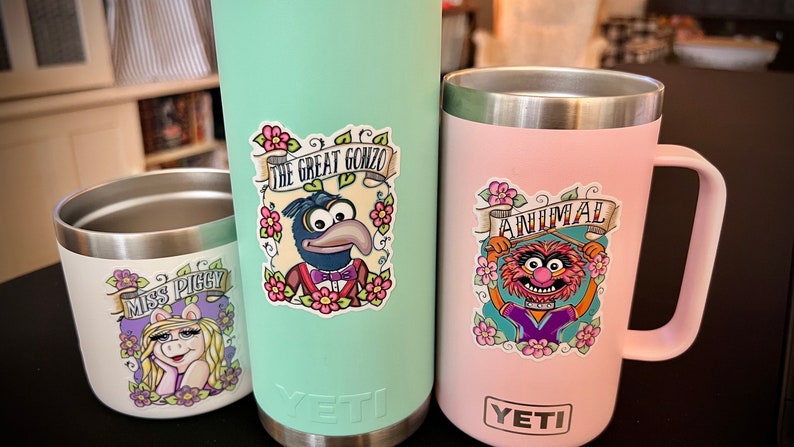 May include: Three stainless steel tumblers with colorful Muppet character stickers. The tumblers are in mint green, pink, and white. The stickers feature Miss Piggy, Animal, and Gonzo. The text on the stickers reads "Miss Piggy", "Animal", and "The Great Gonzo".