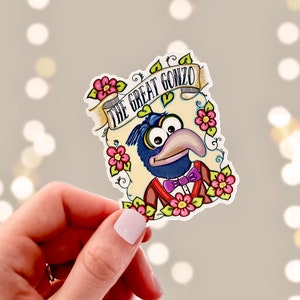 May include: A sticker featuring a cartoon illustration of Gonzo the Great from The Muppets. Gonzo is wearing a blue suit with a bow tie and is surrounded by flowers. The text "The Great Gonzo" is written in a banner above him.
