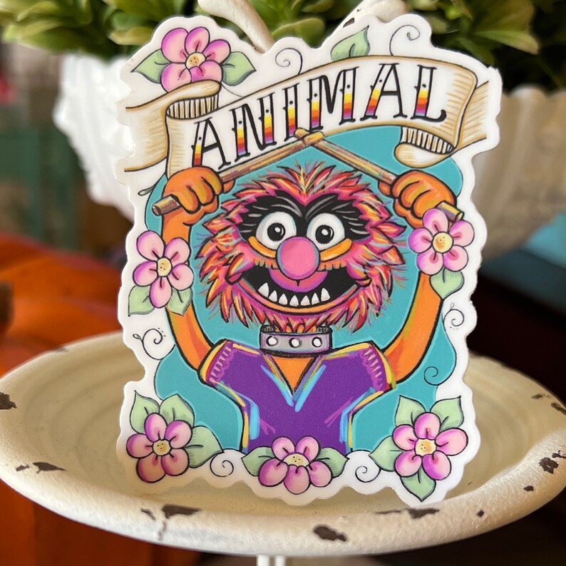 May include: A sticker featuring Animal from the Muppets, a red and orange furry character with a purple shirt, holding drumsticks and a banner that says "ANIMAL". The sticker is surrounded by pink flowers.