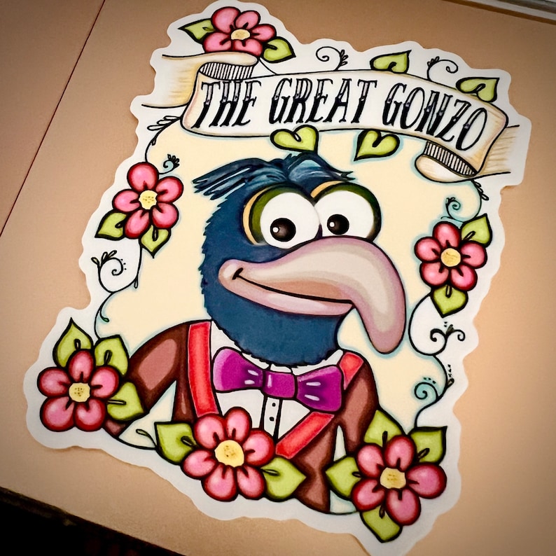 May include: A cartoon illustration of Gonzo the Great, a blue Muppet with a long nose, wearing a brown suit with a red vest and a purple bow tie. The illustration is in a vintage tattoo style with flowers and vines.