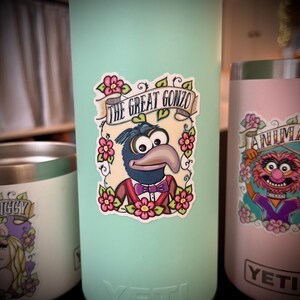 May include: A sticker featuring a cartoon illustration of Gonzo the Great from the Muppets. The sticker is on a light blue Yeti tumbler. The sticker features Gonzo in a blue suit with a purple bow tie and a floral design around him. The text "The Great Gonzo" is in a banner above him.