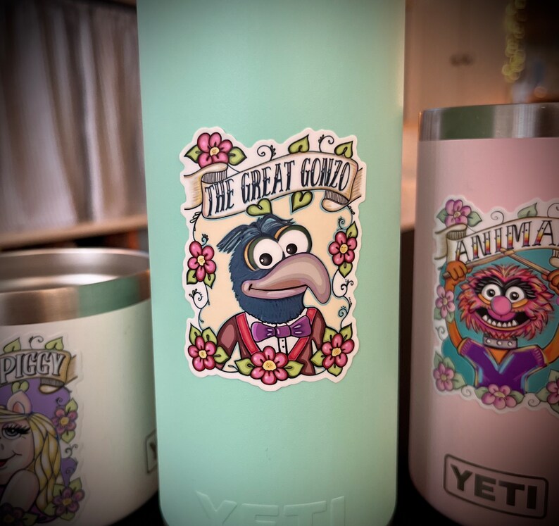 May include: A sticker featuring a cartoon illustration of Gonzo the Great from the Muppets. The sticker is on a light blue Yeti tumbler. The sticker features Gonzo in a blue suit with a purple bow tie and a floral design around him. The text "The Great Gonzo" is in a banner above him.