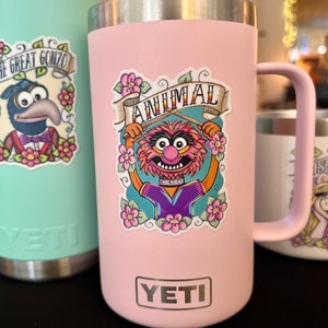 May include: Pink Yeti tumbler with a sticker of Animal from The Muppets playing the drums. The sticker has the words "Animal" and "The Muppets" on it. The tumbler has the Yeti logo on the bottom.