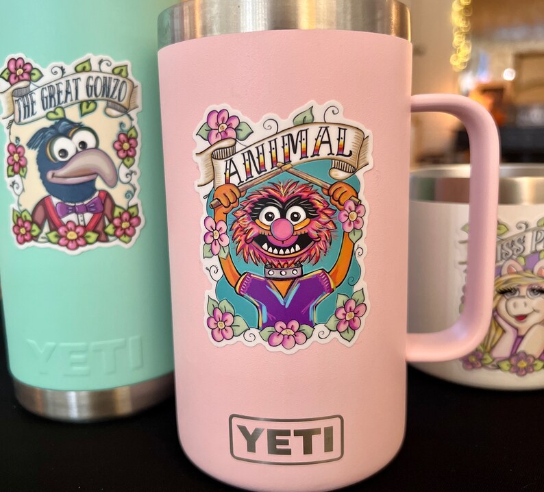 May include: Pink Yeti tumbler with a sticker of Animal from The Muppets playing the drums. The sticker has the words "Animal" and "The Muppets" on it. The tumbler has the Yeti logo on the bottom.