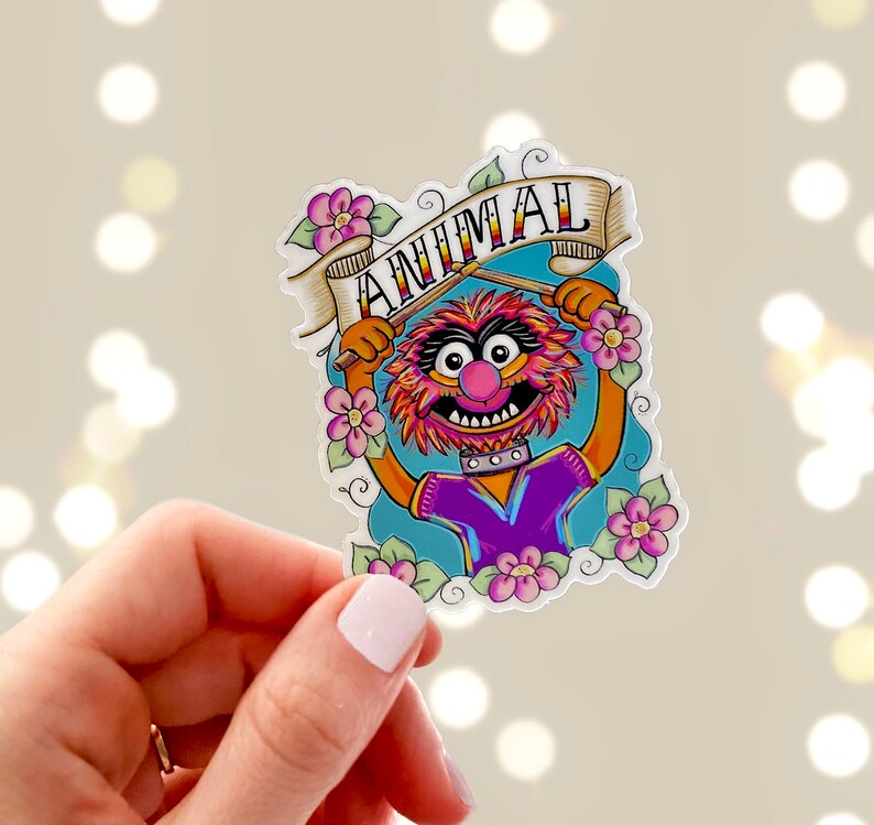 May include: A sticker featuring Animal from The Muppets, a red and orange furry character with a purple shirt, holding drumsticks and a banner that says "ANIMAL". The sticker is surrounded by pink flowers.