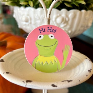 Puede incluir: Una pegatina circular rosa con un personaje de dibujos animados de rana verde que saluda. La rana lleva un collar amarillo y tiene el texto "Hi Ho!" encima.