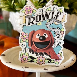 May include: A sticker featuring a cartoon dog playing a keyboard with musical notes and flowers. The dog is brown with a black nose and a big smile. The text "ROWLF" is written in black letters on a white banner.
