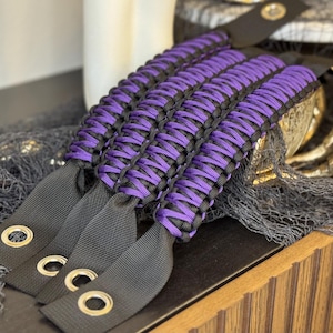 May include: Purple and black paracord wrist straps with metal rings. The straps are woven together in a complex pattern.